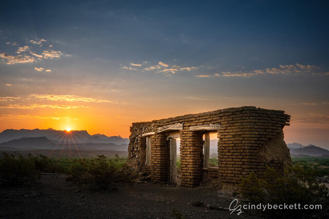 The sun rises over the distant mountains of the Chisos Basin area with the historic adobe structure known as Dorgan farm sitting on the foreground mesa.