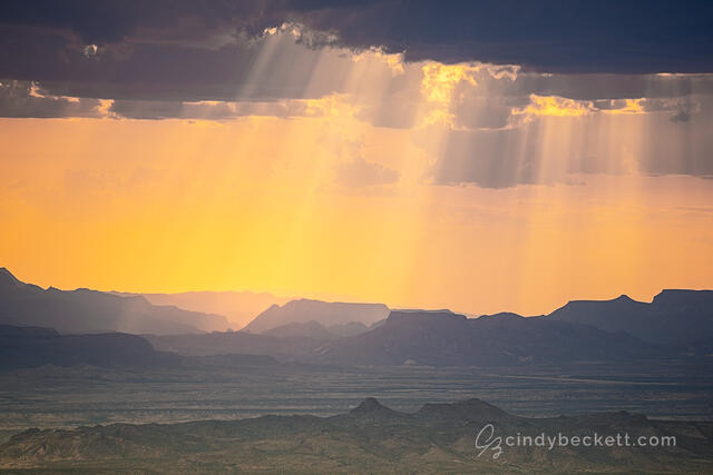 Sun rays shine through thunderstorm clouds as warm afternoon light bathes the scene over Santa Elena Canyon and the Burro Mesa of Big Bend National Park.