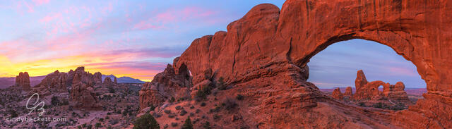 Panorama of Turret Arch through the north window with the south window and other hoodoos in the scene with colorful dawn light and clouds.
