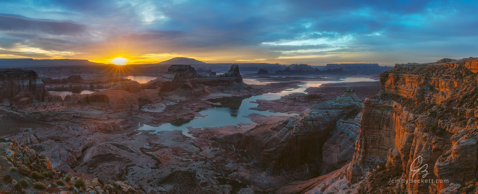 Sun beams breaking over the distant buttes and mesas of Glen Canyon cast warm light on the scene around Gunsight Butte and the waters of Lake Powell. 